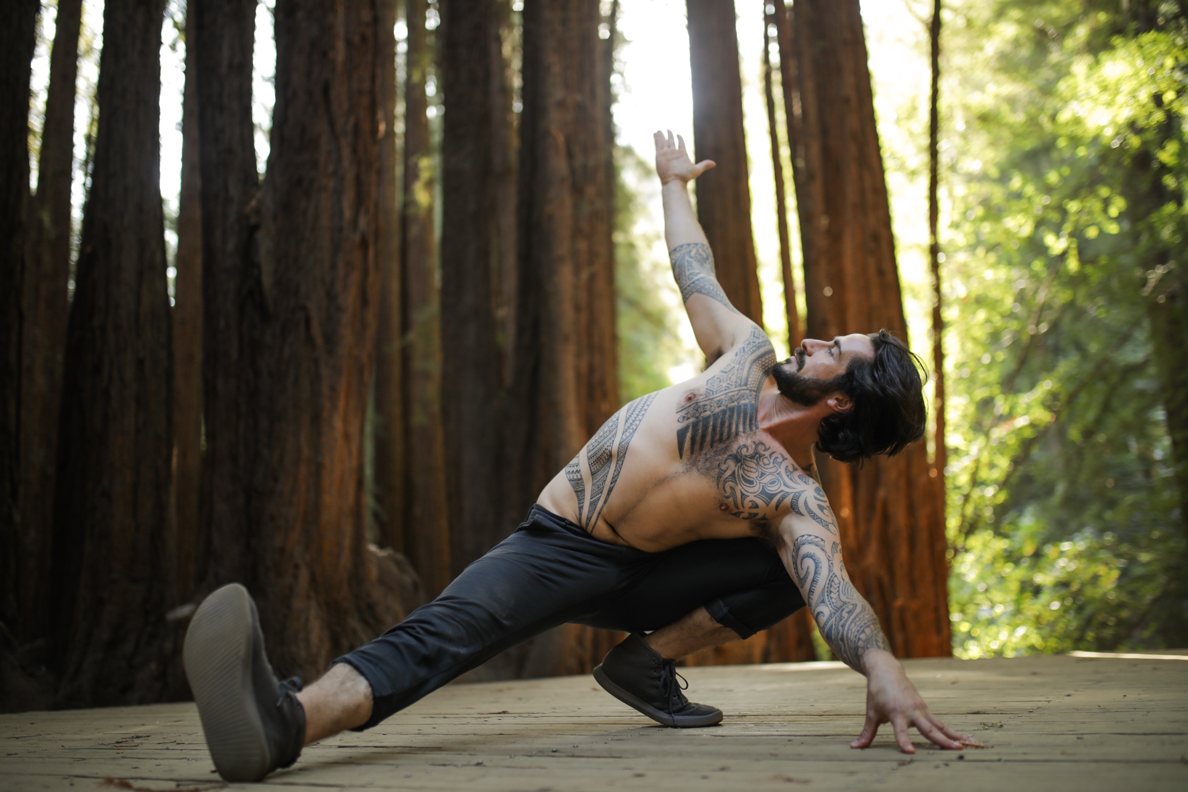 Peter in a deep side-angle lunge on a wood deck in a redwood grove, arm reaching to the sky.